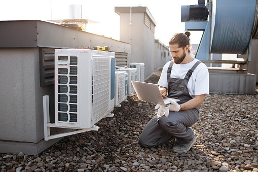 Busy factory worker wearing uniform squatting and using wireless laptop on open air terrace. Caucasian bearded man servicing air conditioners and looking at information on internet on factory.