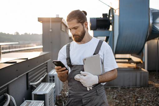 Focused bearded master dressed in overalls standing and holding tablet while using digital smartphone on fresh air. Caucasian skilled man using gadgets and maintaining equipment on roof of factory.