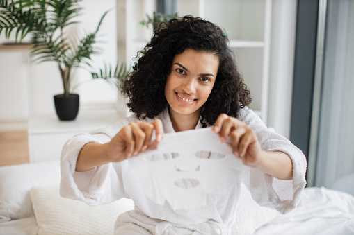 Happy african lady benefiting convenient beauty product of face skincare. Relaxed multiracial female in after-shower outfit using tissue face mask while enjoying day spa at home during weekend.