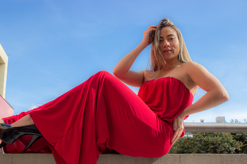 beautiful young woman wearing a red dress sitting in a wall posing with city and sky in background