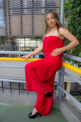 beautiful young woman wearing red dress posing in a bridge with building in the background
