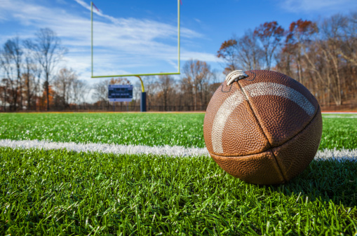 An american football sits on a turf football field with the goal post and score board in the background.