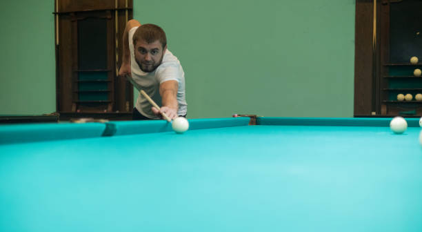 istock Cute man aiming a cue at a billiard ball. In the evening, a man relaxes and relaxes while playing billiards. A man hits the ball with a cue, aiming for a pocket on a billiard table. 1876512640