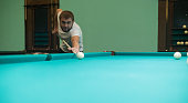Cute man aiming a cue at a billiard ball. In the evening, a man relaxes and relaxes while playing billiards. A man hits the ball with a cue, aiming for a pocket on a billiard table.