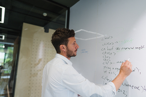 Photo of a an engineer writing a programming code on a transparent wipe board and thinking of a solution for his work-related problem