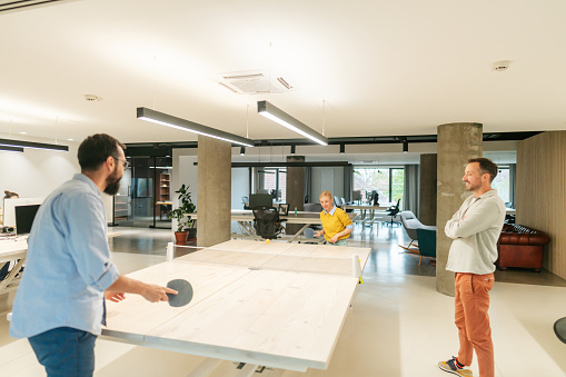 Photo of couple of colleagues having fun during work hours in the office: playing table tennis (ping pong). Their colleagues are cheering for them.