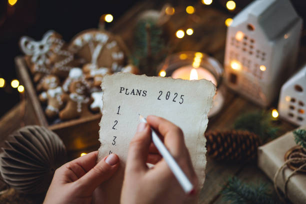 writing new year resolutions 2025 by hand in december on new year's eve. plan, goals, ideas, projects. cozy home holiday atmosphere, christmas lights, festive decor, homemade gingerbread cookies - determination new years eve list aspirations photos et images de collection