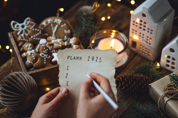 writing new year resolutions 2025 by hand in december on new year's eve. plan, goals, ideas, projects. cozy home holiday atmosphere, christmas lights, festive decor, homemade gingerbread cookies - determination new years eve list aspirations photos et images de collection