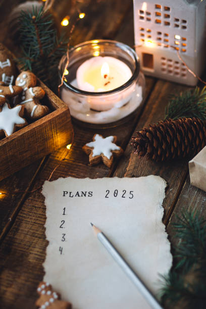 writing new year resolutions 2025 by hand in december on new year's eve. plan, goals, ideas, projects. cozy home holiday atmosphere, christmas lights, festive decor, homemade gingerbread cookies - determination new years eve aspirations letter foto e immagini stock