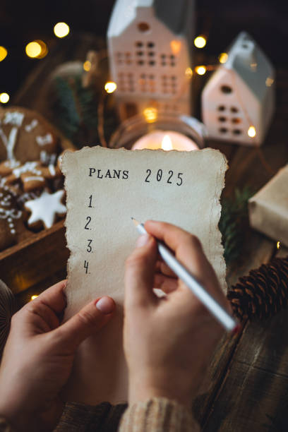 writing new year resolutions 2025 by hand in december on new year's eve. plan, goals, ideas, projects. cozy home holiday atmosphere, christmas lights, festive decor, homemade gingerbread cookies - determination new years eve list aspirations photos et images de collection