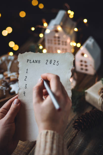 writing new year resolutions 2025 by hand in december on new year's eve. plan, goals, ideas, projects. cozy home holiday atmosphere, christmas lights, festive decor, homemade gingerbread cookies - determination new years eve list aspirations 뉴스 사진 이미지