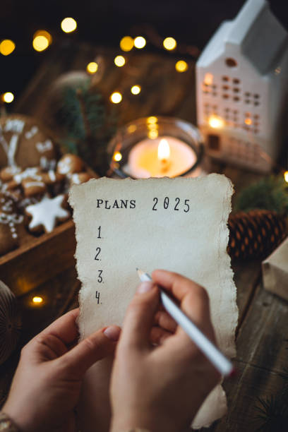 writing new year resolutions 2025 by hand in december on new year's eve. plan, goals, ideas, projects. cozy home holiday atmosphere, christmas lights, festive decor, homemade gingerbread cookies - determination new years eve list aspirations photos et images de collection