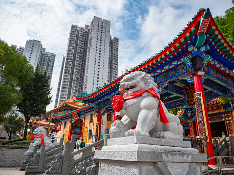 November 13, 2023. Wong Tai Sin Temple - Wong Tai Sin area, Hong Kong. Stone color statue of lion dog in front of Wong Tai Sin temple building decorated with red color ribbon during the day