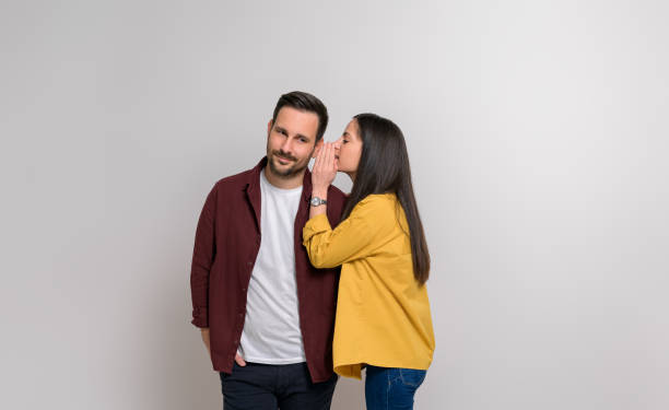 istock Young girlfriend whispering secret in handsome boyfriend's ear while standing on white background 1876720132