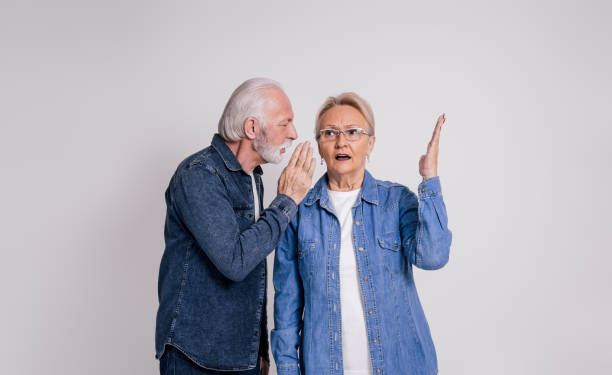 istock Senior husband whispering gossip in shocked wife's ear gesturing while standing on white background 1876725297