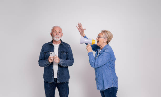 istock Senior woman shouting over megaphone at shocked husband using smart phone over white background 1876725305