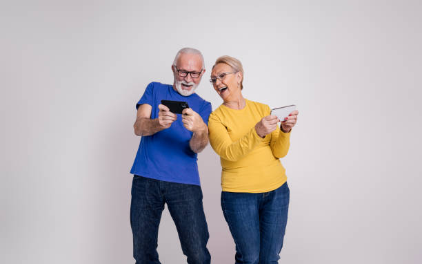 istock Senior couple laughing and playing video game over mobile phone while standing on white background 1876728985