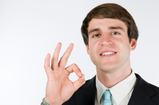 Head and shoulder came of a young man in black suit against white background showing the OK sign in the camera