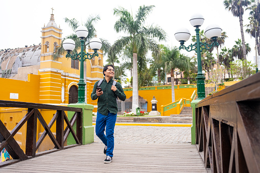 Wide full length view of cheerful  young Hispanic man taking a stroll while sightseeing in Puente de los Suspiros, Iglesia la Ermita, Barranco, Lima, Peru