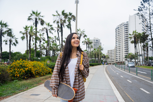 Mid-shot front view of cheerful young Latin woman holding a skateboard while walking in streets of Miraflores, Peru
