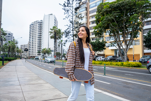Mid-shot view of young Hispanic woman holding skateboard while walking in streets of Miraflores, Peru