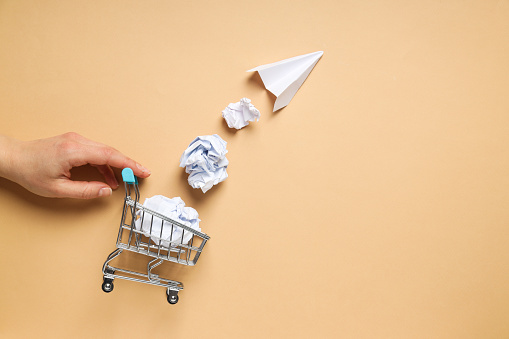 Shop cart, paper balls, paper plane and hand on beige background