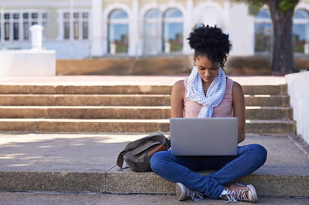 She wants to do well in her Degree An ethnic woman sitting on the steps of her college doing research on her laptop legs crossed at knee stock pictures, royalty-free photos & images