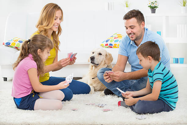 Cheerful Family Playing Cards. Cheerful young family with two children having fun together, sitting on carpet at home and playing cards. Their pet, dog Retriever enjoy with them. family playing card game stock pictures, royalty-free photos & images