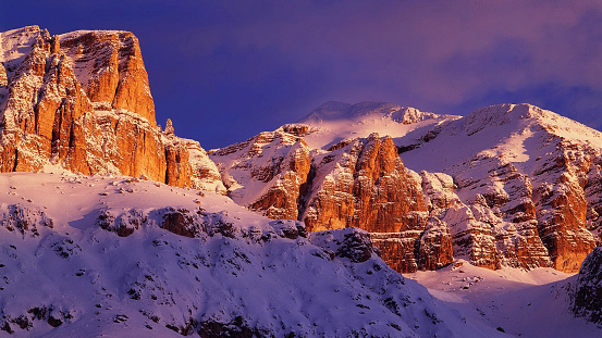 Witness the Dolomites' magic hour as Sella Group's snow-kissed peaks ignite with vibrant hues. A symphony of Purple, Pink, and Orange skies dances amidst wispy clouds, casting an Alpenglow spell on the rugged landscape.