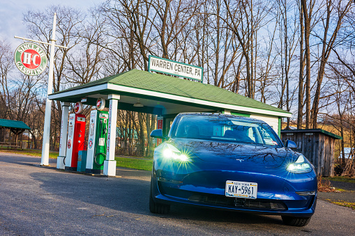 Warren Center, PA, USA - Dec 22, 2023: A blue Tesla Model 3, an electric vehicle, is photographed near a vintage gas station located in a rural town. Are EVs a fad or here to stay?