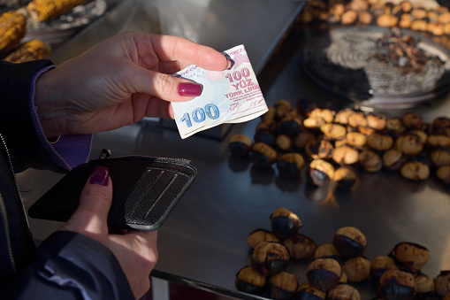 Istanbul, Turkey - December 10, 2023: A woman buys street food in Istanbul. Hand with 100 Turkish lira banknot close-up. Roasted chestnuts and corn. Turkey