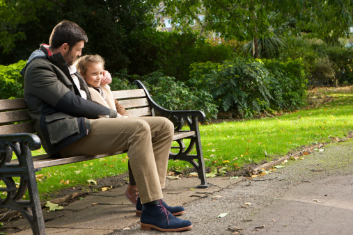 A father and daughter sitting on a park bench as dad gently squeezes his little girl's cheek. 