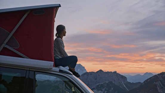 Captured in a Serene Moment,a Side View Reveals a Woman Enjoying the Scenic Sunset Over Mountains While Seated Outside a Rooftop Tent on a Camper Van. The Scene Harmonizes the Tranquility of the Evening with the Breathtaking Beauty of the Mountainous Landscape