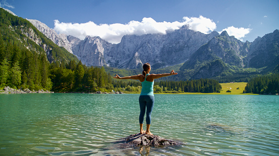 In a Scene of Serenity and Strength,a Sporty Woman Stands with Arms Outstretched,Meditating on a Rock Surrounded by a Rippled Lake Against the Backdrop of Majestic Rocky Mountains. The Image Captures the Harmonious Blend of Mindfulness and the Raw Beauty of Nature