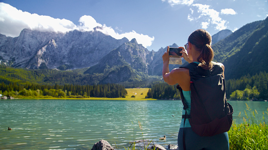 Amidst the Beauty of Summer Vacations,a Female Hiker is Seen Capturing the Scenic View of a Lake and Rocky Mountains Through Her Mobile Phone. The Image Encapsulates the Modern Traveler's Blend of Technology and Nature Appreciation,Creating a Timeless Memory of the Journey