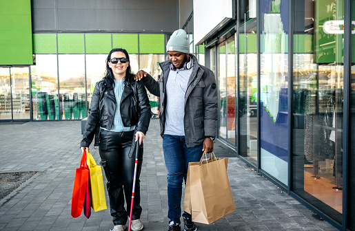 Blind person carrying shopping bags and walking with friend in front of shop window