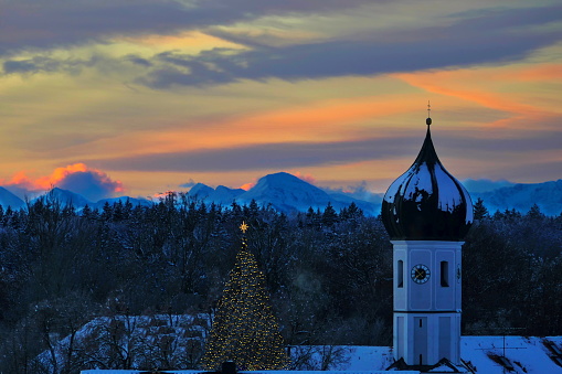Bavaria, Germany - Cristmas 2023. Panoramic view of our village at sunrise with characteristic Bavarian white and blue cloudy sky and the alps at the background. A new  illuminated Christmas tree can be seen next to the church.