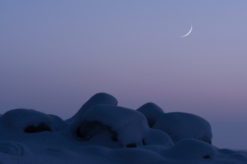 Winter landscape at twilight. Crescent new moon in the sky behind snow covered boulders.