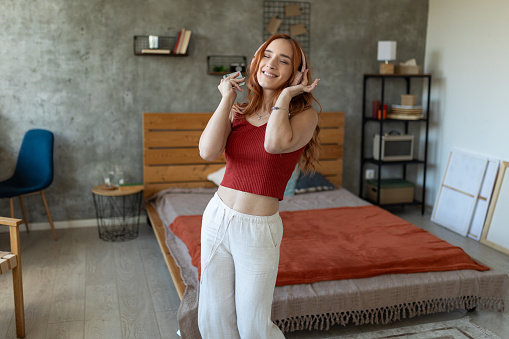 Beautiful redhead woman dancing while listening music on wireless headphones in bedroom at home.