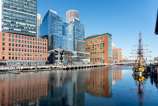 Buildings along the Boston waterfront are reflected in Boston Harbor. On the right is a replica 18th century British merchant vessel  that participates in the 250th anniversary celebration of the Boston Tea Party.