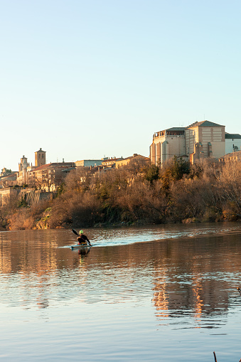 On the Duero River as it passes through Tordesillas, Castilla y Leon-Spain. At dusk of an autumn day with the golden sunlight reflecting on the buildings of the town, a young canoeist is training. Leisure activity on a canoe in an autumn sunset, a young canoeist is on the river paddling.