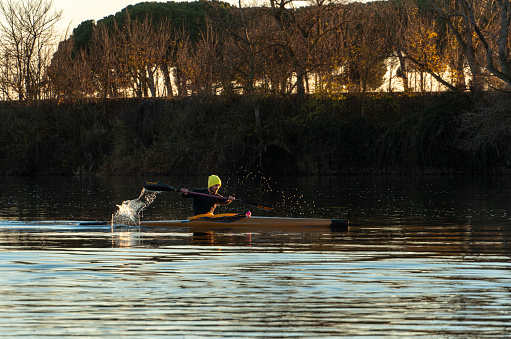 On the Duero River as it passes through Tordesillas, Castilla y Leon-Spain. At dusk of an autumn day with the golden sunlight reflecting on the trees of the riverbank, a young canoeist is training. Leisure activity on a canoe in an autumn sunset, a young canoeist is on the river paddling.