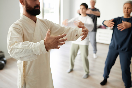 Cropped shot of bearded male qigong master teaching qigong tree pose, students repeating standing meditation exercise in blurred background