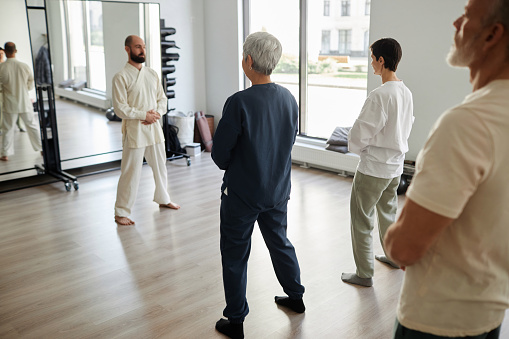 Back view at group of senior people standing in gym and meditating while qigong instructor observing