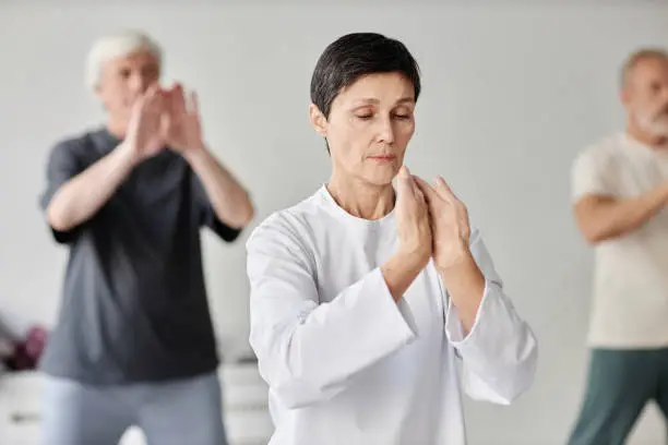 Medium shot of brunette senior Caucasian lady concentrating on qigong exercise and moving her hands, male class participants exercising in blurred background