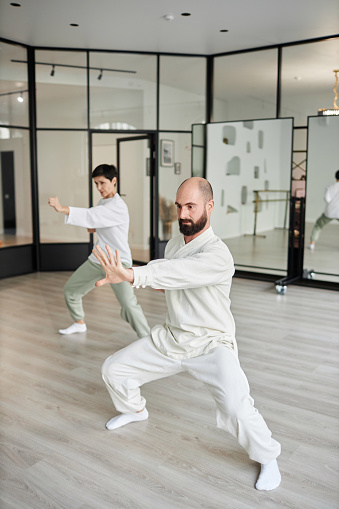 Vertical full length shot of qigong master and woman student practicing energy punch pose in modern gym while standing in half-squat with hands outstretched