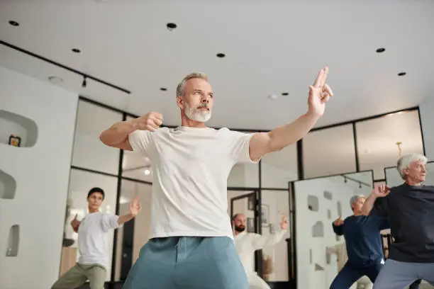 Low angle view of senior people exercising in modern gym, focus on concentrated elderly man in qigong archer pose