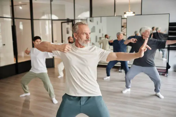 Side view of elderly people in gym, focus on bearded senior man posing as archer shooting during qigong lesson in gym