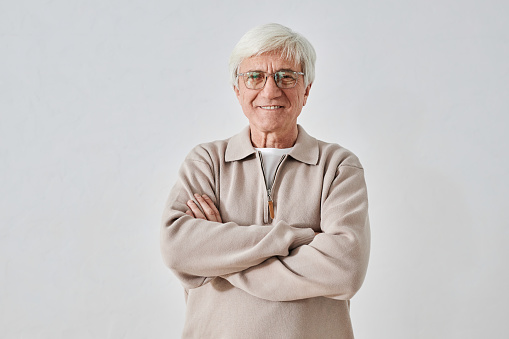Waist up portrait of smiling white-haired senior man wearing beige sweater and glasses standing with arms crossed on white background while looking at camera