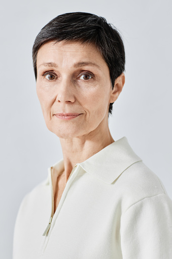 Vertical close up shot of calm Caucasian dark-haired senior woman in white sweater standing body half-turned and looking at camera